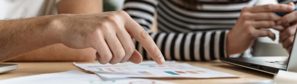 Duas pessoas sorridentes analisam gráficos coloridos em uma folha de papel sobre a mesa. Uma delas aponta para os dados enquanto a outra segura uma caneta, com um notebook parcialmente visível ao lado. A cena transmite aprendizado colaborativo, planejamento financeiro e análise de informações de forma descontraída e acessível.