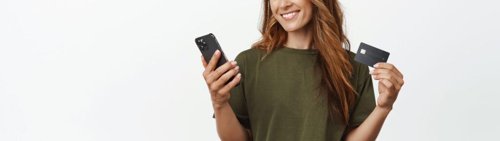 Mulher jovem sorridente, usando camiseta verde e jeans, segura um cartão de crédito em uma mão e um smartphone na outra. Ela olha para o celular com expressão satisfeita, em frente a um fundo branco. A imagem representa facilidade no uso de tecnologia para controle financeiro ou realização de pagamentos digitais.