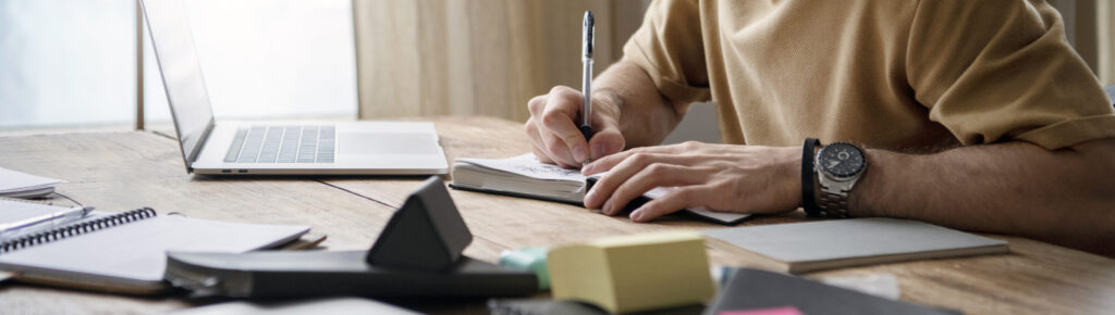 Homem jovem usando fones de ouvido e óculos estuda em uma mesa de madeira, escrevendo em um caderno enquanto consulta um notebook. Há vários materiais de papelaria sobre a mesa, como post-its, cadernos e canetas. A luz natural entra pela janela ao fundo. A cena transmite concentração, aprendizado e organização, associada ao estudo ou planejamento financeiro.