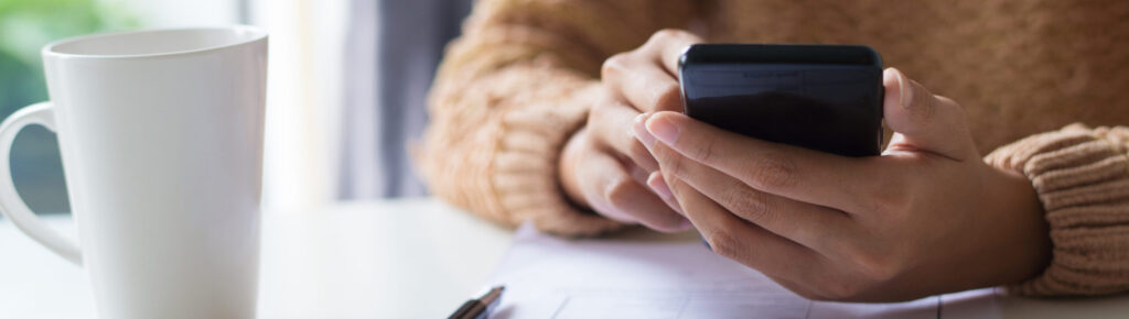 Pessoa usando um smartphone enquanto analisa um formulário sobre uma mesa; ao lado, há uma caneta e uma xícara branca. A pessoa veste um suéter marrom.