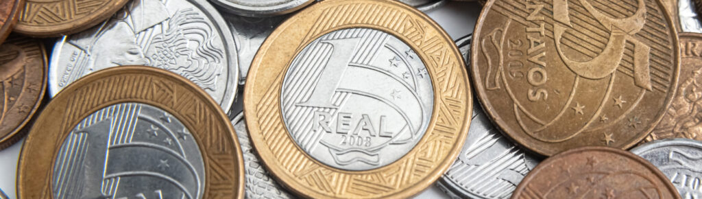 Várias moedas brasileiras de diferentes valores espalhadas, incluindo peças de 1 real, 25 centavos, 10 centavos e 5 centavos, em diferentes estados de conservação.