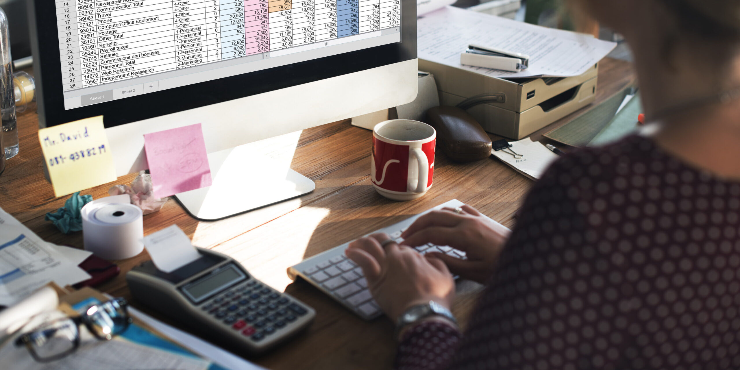Mulher analisando planilha financeira no computador, com dados orçamentários coloridos e calculadora sobre a mesa, representando organização e controle financeiro.