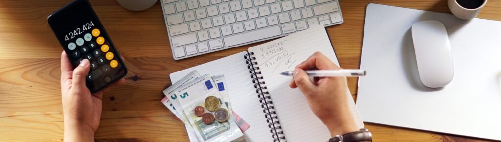 Pessoa fazendo cálculos com celular e anotando em caderno, com dinheiro em espécie sobre a mesa, representando controle financeiro pessoal, orçamento doméstico e planejamento financeiro em home office.