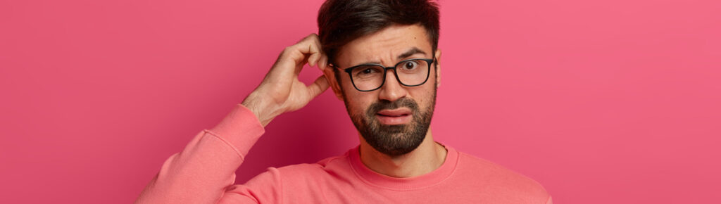 Homem jovem de expressão confusa coça a cabeça, vestindo suéter rosa e óculos, em frente a fundo rosa. A imagem transmite dúvida ou indecisão.