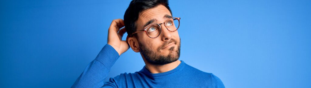 Homem jovem com barba, usando óculos e suéter azul, coçando a cabeça com expressão de dúvida, em frente a um fundo azul.