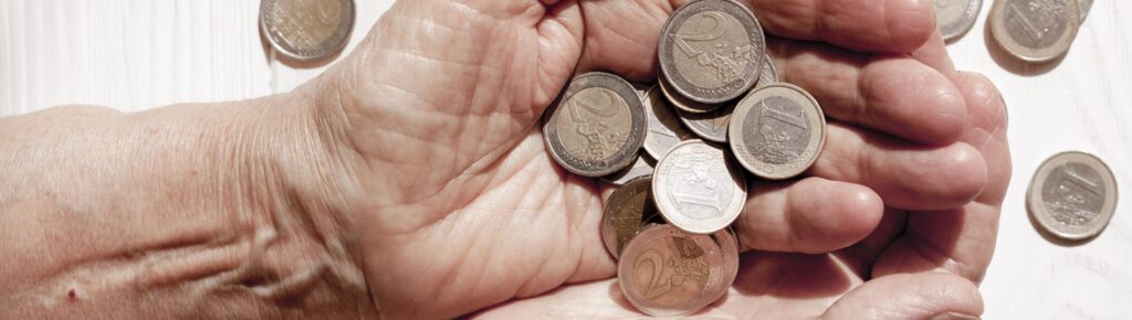 Mãos idosas segurando várias moedas de euro, com algumas espalhadas sobre uma superfície branca de madeira.
