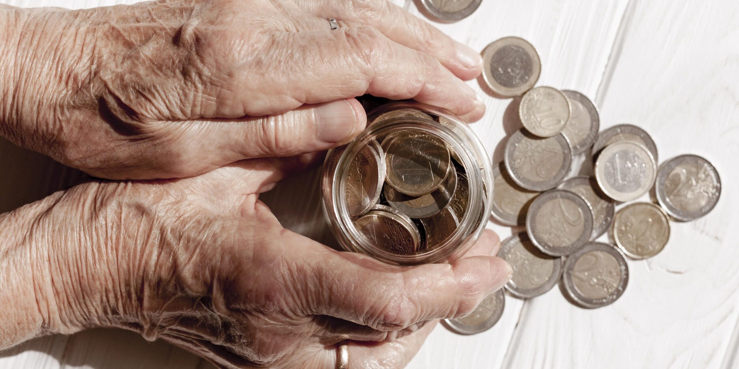 Mãos idosas segurando um pequeno frasco de vidro cheio de moedas, com várias outras moedas espalhadas sobre uma superfície branca de madeira.