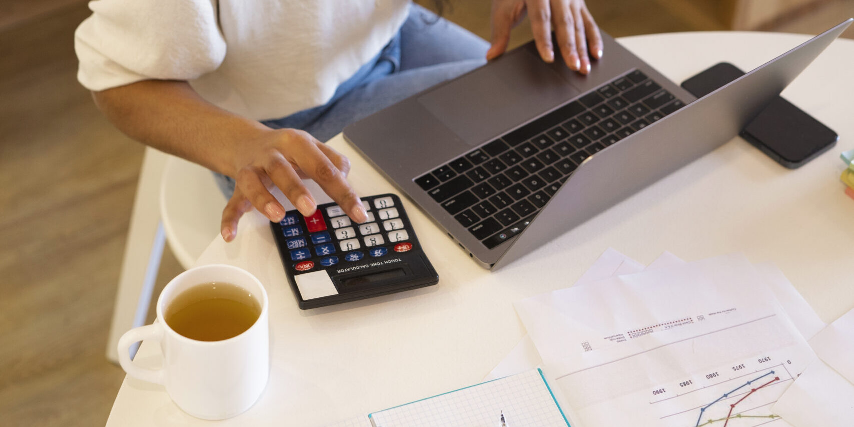 Mulher jovem trabalhando com laptop e calculadora em ambiente de escritório, analisando gráficos financeiros e tomando notas, representando gestão financeira e produtividade