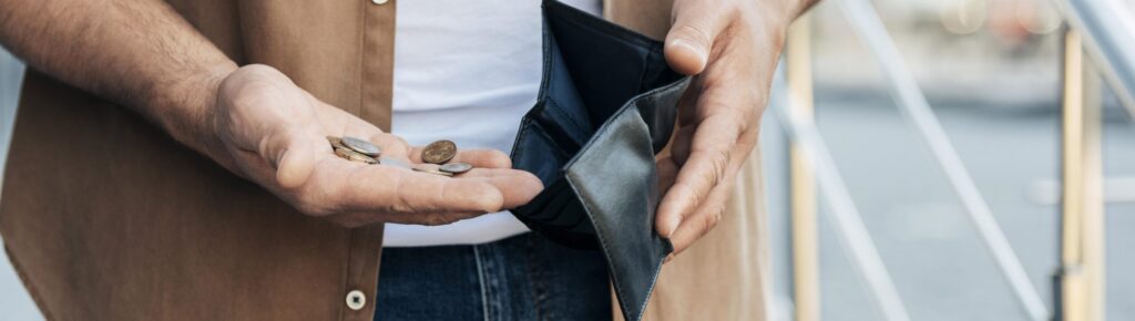 Homem segurando moedas em uma mão e mostrando uma carteira vazia com a outra, simbolizando dificuldades financeiras ou falta de recursos para poupar.