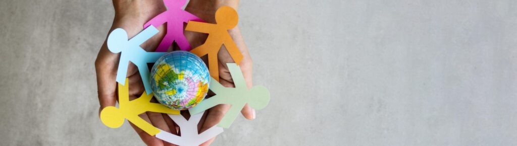 Mãos acolhem uma corrente de figuras humanas coloridas de papel que formam um círculo ao redor de um pequeno globo terrestre, simbolizando união, cuidado e amor entre os povos do mundo.