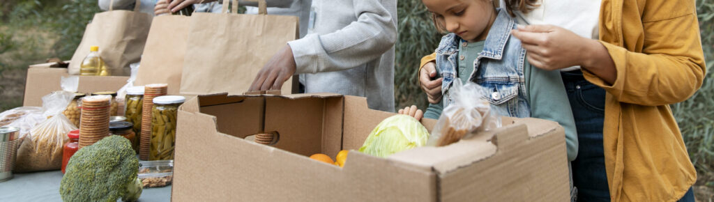 Voluntários organizando doações de alimentos em sacolas e caixas, com presença de mulher e criança recebendo mantimentos em ação solidária ao ar livre.