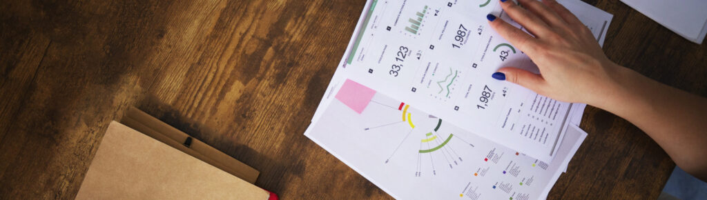 Vista superior de mulher analisando relatórios financeiros com gráficos e usando calculadora no celular, representando planejamento financeiro e controle de dados econômicos.