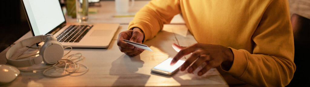 Homem sorridente usando cartão de crédito e smartphone para pagamento online enquanto trabalha à noite no escritório.