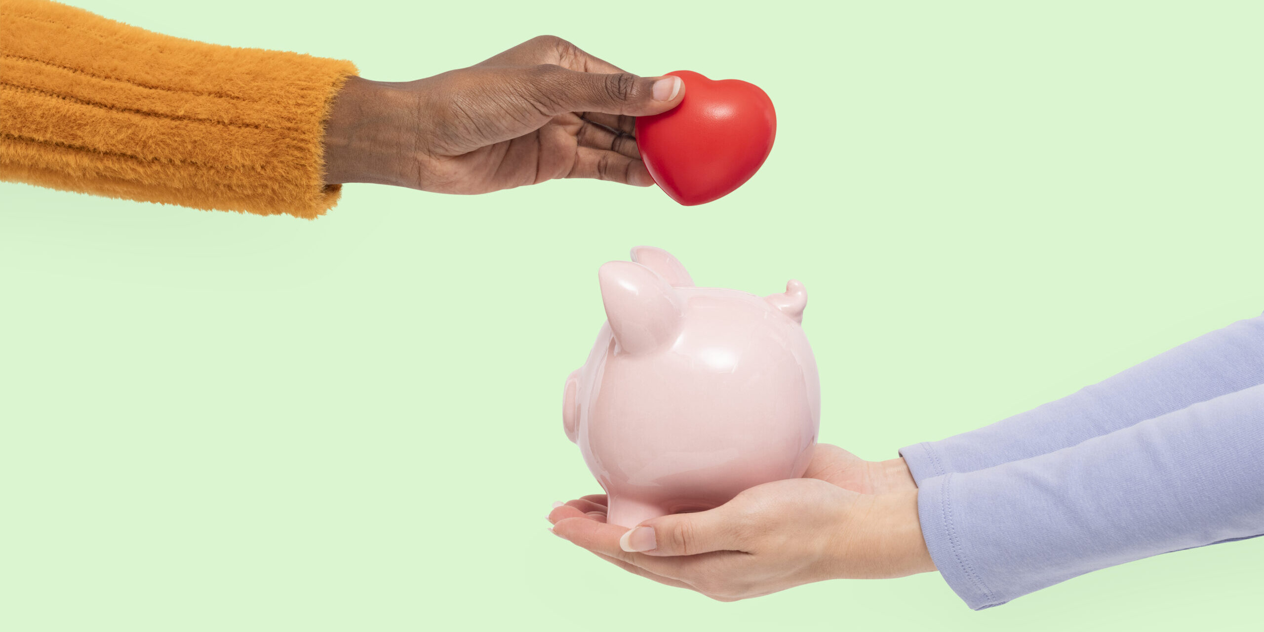 Mão segurando coração vermelho sobre cofrinho rosa em formato de porco, simbolizando poupança, finanças solidárias, investimento com propósito e economia colaborativa.