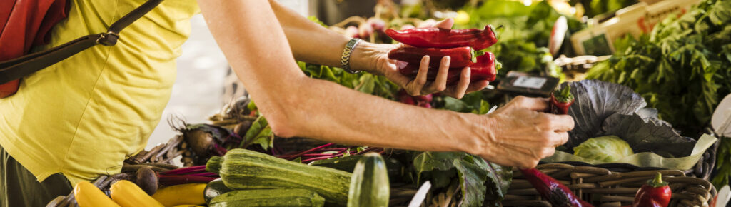 Pessoa escolhendo pimentões vermelhos frescos em feira livre com bancadas cheias de tomates, abobrinhas e hortaliças, representando consumo local, agricultura familiar e alimentação saudável.