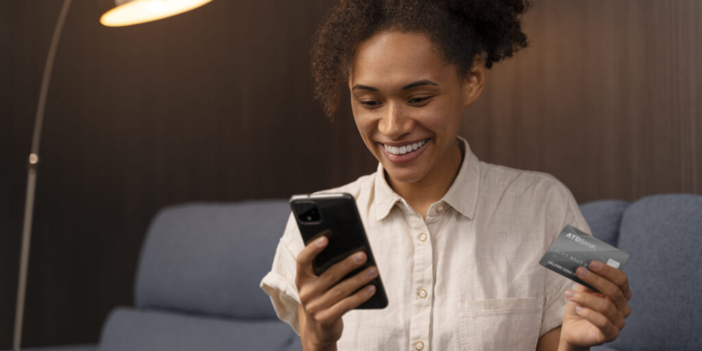 Mulher jovem sorridente usando smartphone e segurando cartão de crédito no sofá, representando compras online e gestão financeira digital.