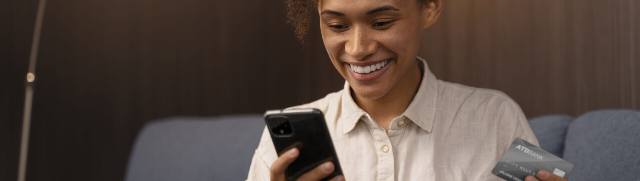 Mulher jovem sorridente usando smartphone e segurando cartão de crédito no sofá, representando compras online e gestão financeira digital.