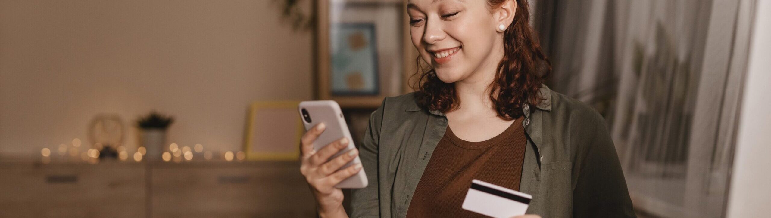 Mulher sorridente usando smartphone e segurando cartão de crédito em casa, representando compras online e controle financeiro digital.