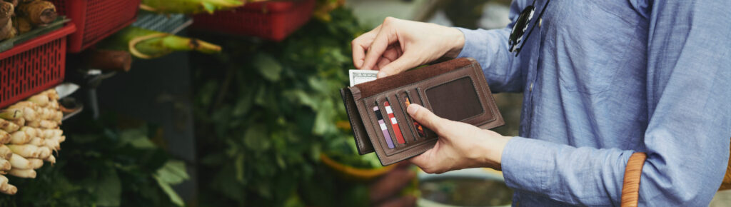 Pessoa pagando compras em feira livre com dinheiro em carteira, segurando cesta de palha cheia de verduras frescas, representando consumo consciente, economia local e apoio a produtores da agricultura familiar.