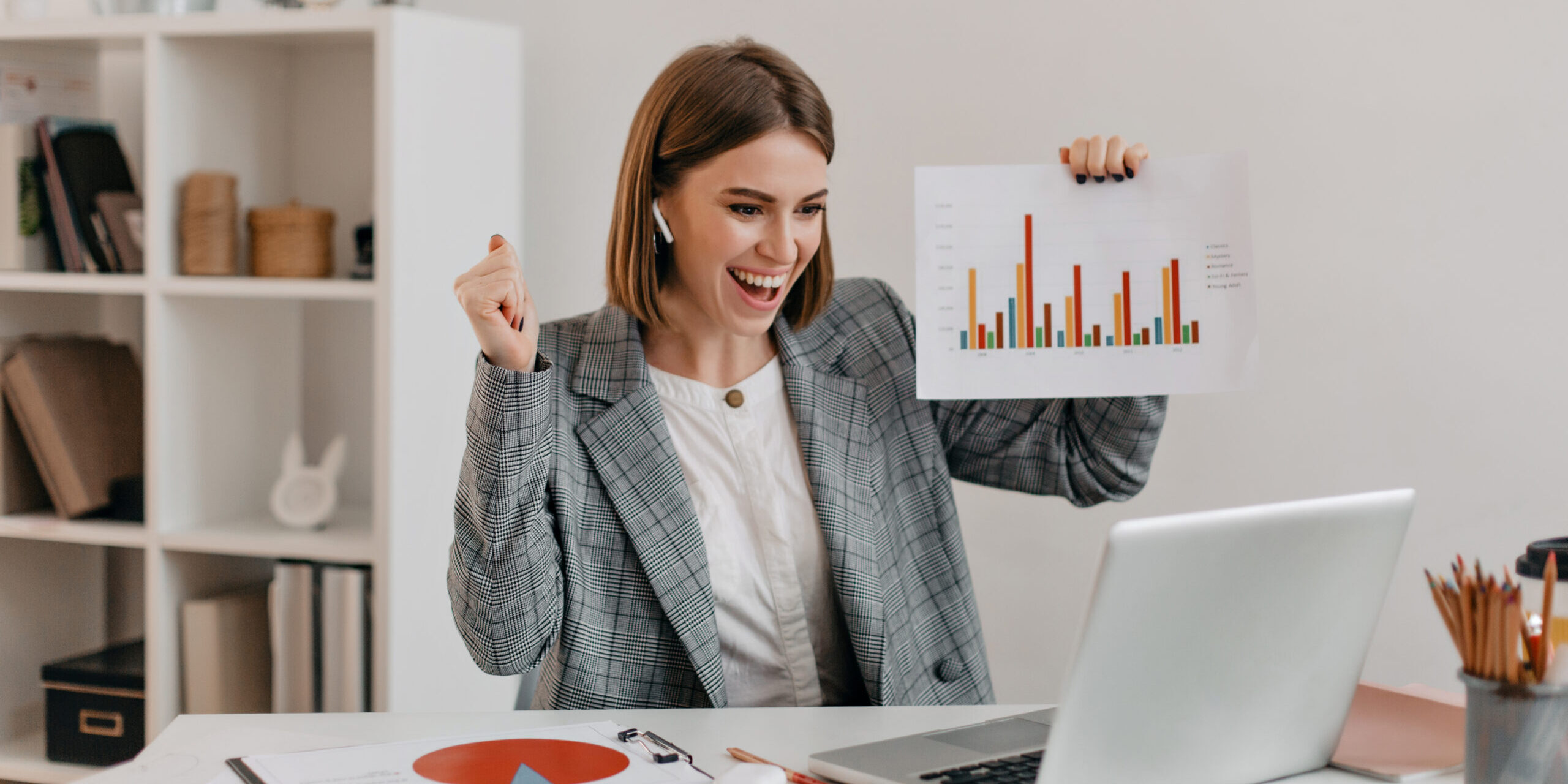 Close-up shot of happy business woman in stylish outfit. Girl in high spirits demonstrates chart via Skype. Mulher de negócios sorridente comemorando resultado positivo em gráfico de barras, sentada em frente ao notebook em ambiente de escritório, representando crescimento financeiro, sucesso profissional e análise de dados.
